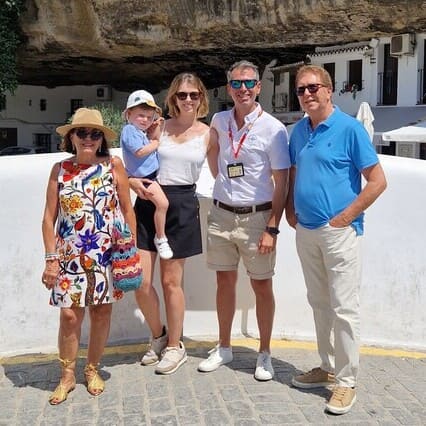A family from Seville and our guide are captured posing on a sunny day during one of our Seville Private Tours. They stand in front of a white balustrade, with a striking rock formation and buildings integrated into the cliff face in the background. This scene reflects the unique charm of Setenil de las Bodegas in Cádiz, Spain, renowned for its homes built into rock overhangs above the Rio Trejo.